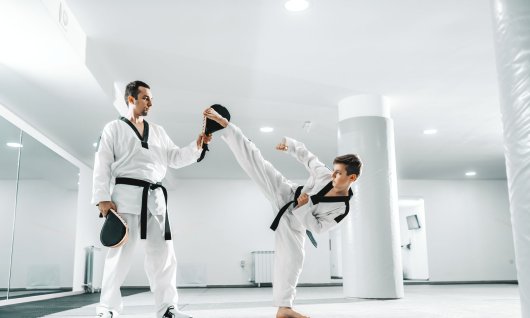 Ein Junge in weißem Taekwondo-Anzug trainiert mit einem Mann mit Schlagpolster Fußtritte