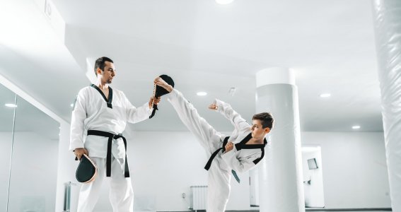 Ein Junge in weißem Taekwondo-Anzug trainiert mit einem Mann mit Schlagpolster Fußtritte