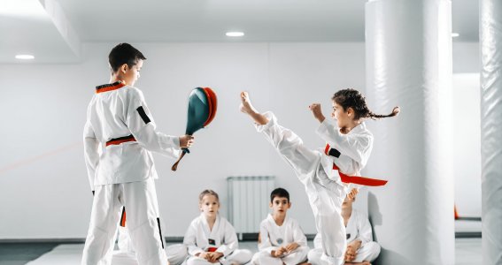 Ein Junge und ein Mädchen in Taekwondo-Anzügen trainieren mit einem Schlagpolster in einem Turnsaal Fußtritte während andere Kinder zuschauen