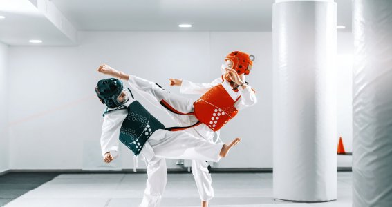 Zwei Jungs mit Helmen und Trainingswesten trainieren in einem leeren, weißen Sportsaal Taekwondo