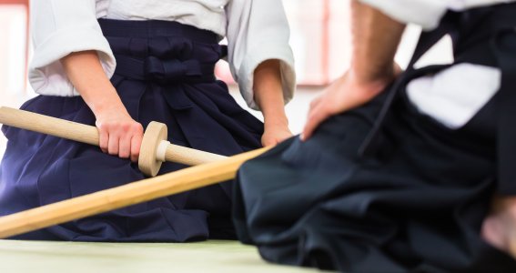 Zwei Menschen in japanischer Trainingskleidung sitzen sich mit Holzstöcken gegenüber