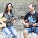 Ein lachender Mann und eine Frau sitzen im Freien und spielen Gitarre