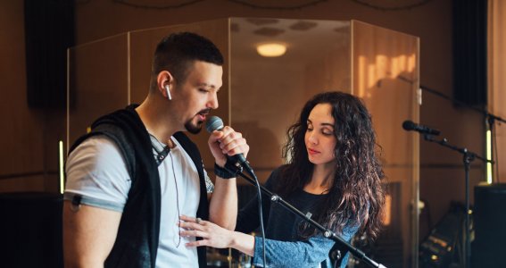 Ein Mann singt in ein Stehmikrofon und eine Frau korrigiert seine Körperhaltung