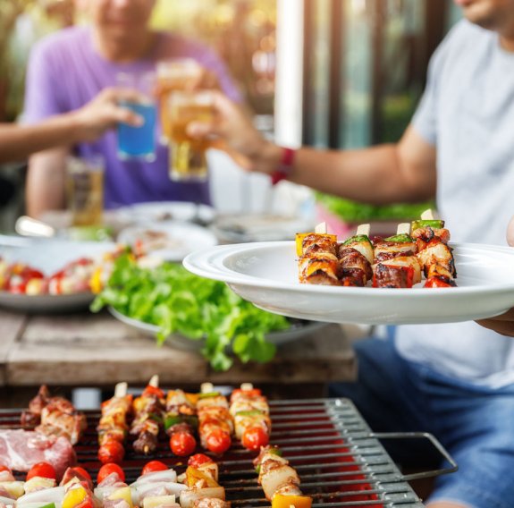 Ein Mann hält einen Teller voller Grillspieße über einem Grill und stoßt mit Freunden an