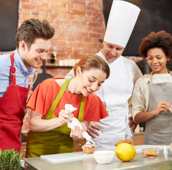 Eine Frau verziert einen Muffin und zwei Personen in Kochschürze und ein Koch schauen ihr zu