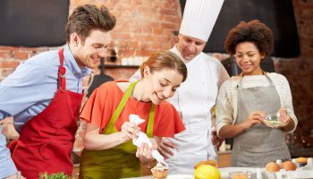 Eine Frau verziert einen Muffin und zwei Personen in Kochschürze und ein Koch schauen ihr zu