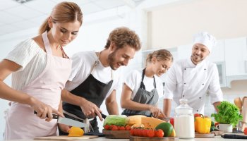 Drei junge Menschen in Kochschürze schneiden Gemüse neben einem Koch