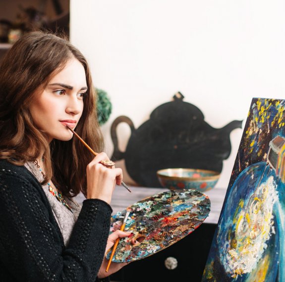 Eine junge Frau steht konzentriert vor einem Gemälde und hält einen Pinsel und eine Farbpalette 