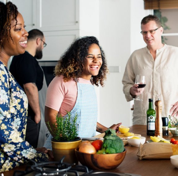 Lachende Menschen in Kochschürzen stehen um einen Küchentisch mit Lebensmitteln 