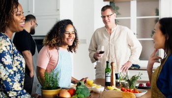Lachende Menschen in Kochschürzen stehen um einen Küchentisch mit Lebensmitteln 