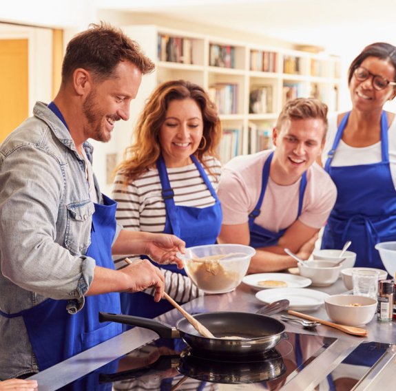 Eine Gruppe lächelnder Erwachsenen in Kochschürze schaut einem Mann beim Kochen zu