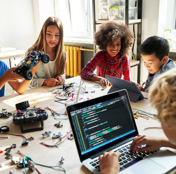 Kindern sitzen um einen Schreibtisch herum und arbeiten am Laptop und bauen ein Elektroauto