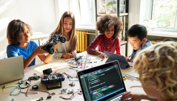 Kindern sitzen um einen Schreibtisch herum und arbeiten am Laptop und bauen ein Elektroauto