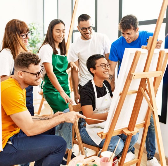 Eine Gruppe lachender Jugendlicher steht und sitzt im Kunstatelier kreisförmig vor einer weißen Leinwand 