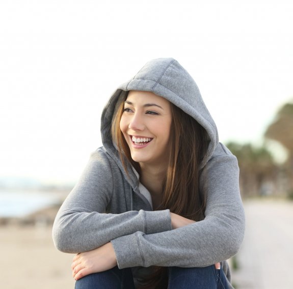 Eine lachende Jugendliche mit grauem Hoodie sitzt am Strand