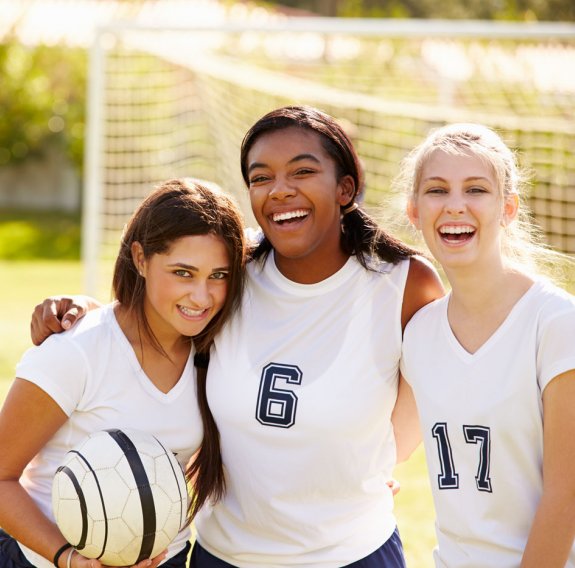 Drei lachende Mädchen halten sich auf dem Fußballfeld in den Armen und halten einen Fußball