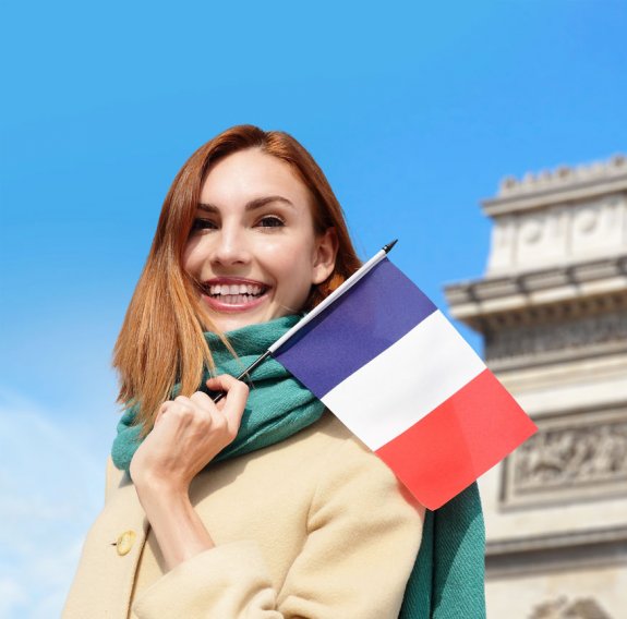 Eine lächelnde Frau im Mantel und Schal hält eine französischer Flagge und steht in Paris vor dem Arc de Triomphe