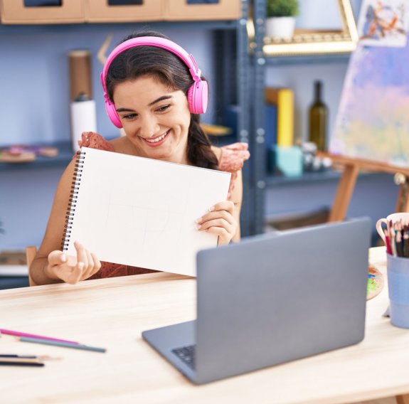 Ein jugendliches Mädchen mit pinken Kopfhörern hält einen Zeichenblock in die Laptop-Kamera und lächelt